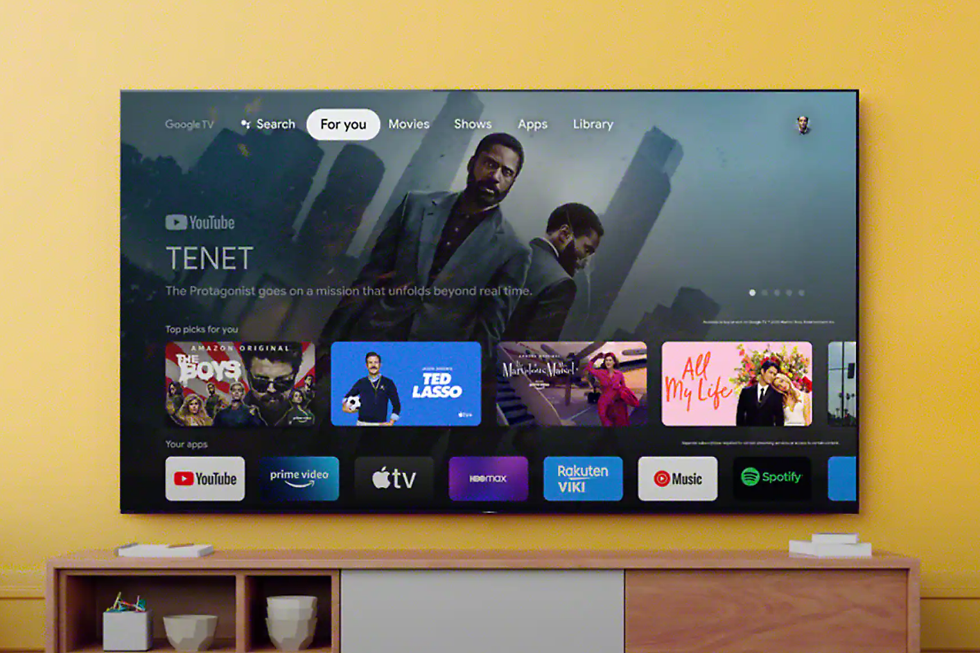 Màn hình TV hiển thị giao diện với điều hướng tìm kiếm và lựa chọn ứng dụng, với một cảnh từ “Tenet” trên màn hình trong nền.