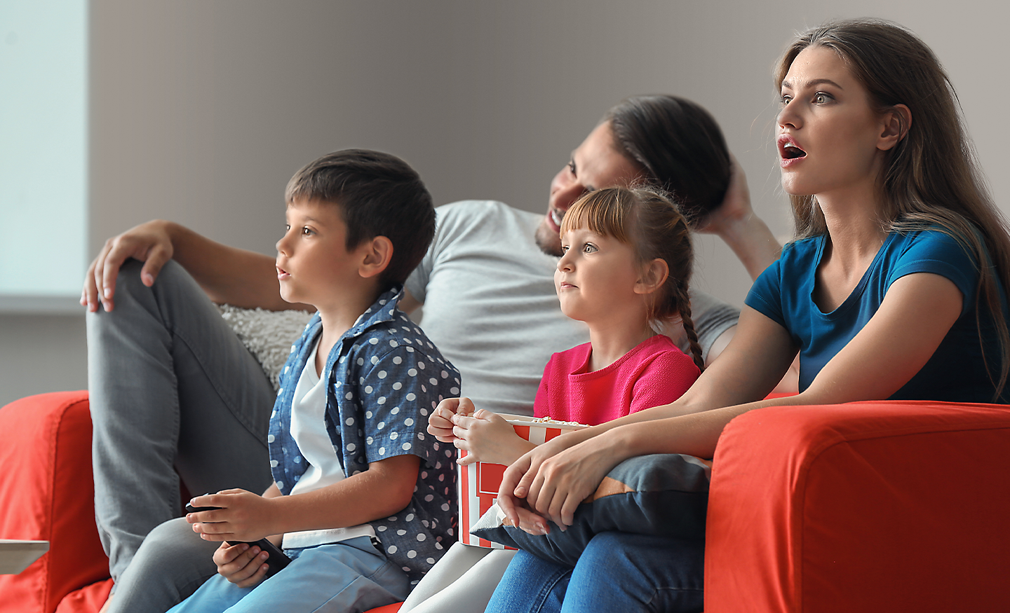 Četvoročlana porodica sedi na kauču, potpuno zaokupljena onim što gledaju na televizoru