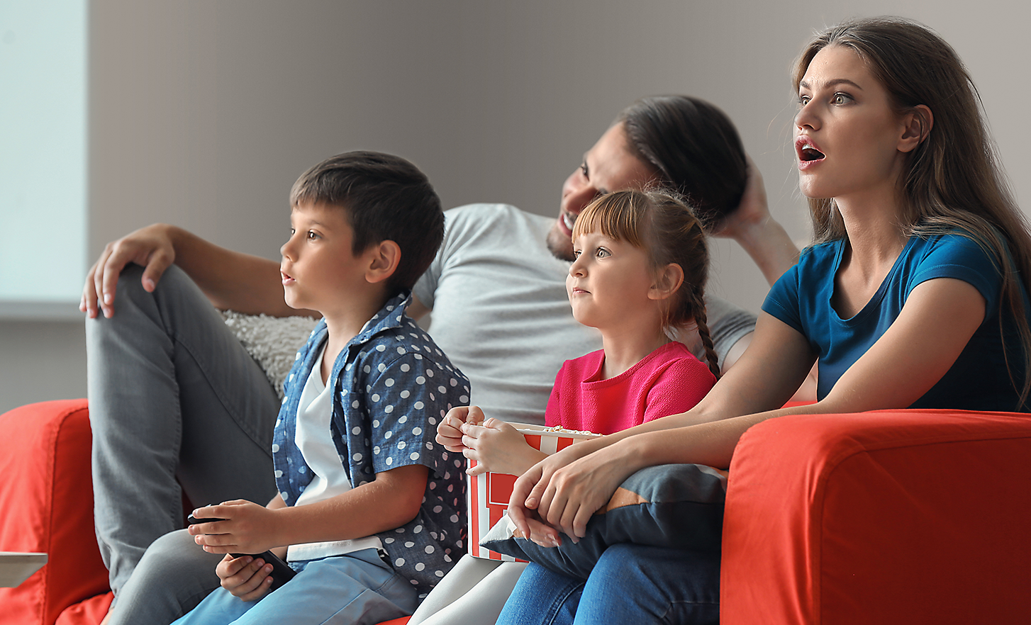 Ant sofos sėdinti keturių asmenų šeima, susižavėjusi tuo, ką žiūri per televizorių