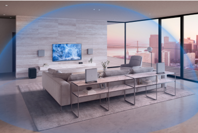 Light brown sofa, table and chair in a living room with wall-mounted TV, surround sound speakers on a display unit and wall, and soundbar on a plinth, with glass wall to the right overlooking a cityscape