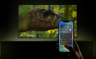 Wall-mounted TV above a plinth with screenshot of dinosaur head, and hand holding a smartphone in the foreground with app on screen