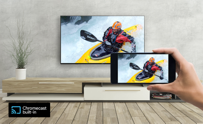 View of living room with wall-mounted TV above a plinth and screenshot of canoeist, and hand holding a smartphone in the foreground with same screenshot