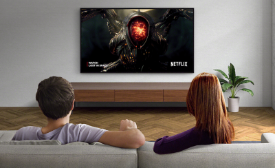 คนสองคนที่นั่งบนโซฟากำลังดูทีวีติดตั้งผนังที่มีภาพหน้าจอ Lost in Space และโลโก้ Netflix ที่ด้านล่างขวา