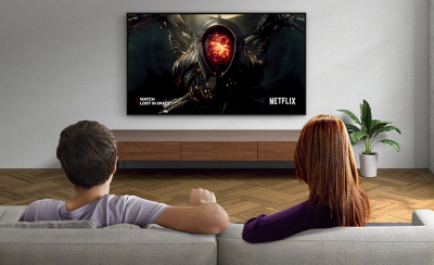 Two people sitting on a sofa watching a wall-mounted TV with screenshot of Lost in Space and Netflix logo bottom right