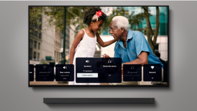 Televizor montat pe perete, deasupra unei bare de sunet, cu imagine cu doua persoane vorbind, cu o scena urbana pe fundal si pictogramele de meniu in partea de jos a ecranului