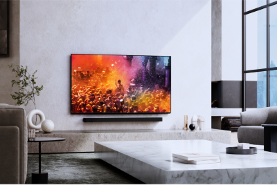 Living room scene with chairs left and right, coffee table centre and wall-mounted TV above soundbar and plinth showing colourful screenshot of crowd at an event