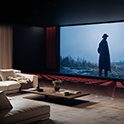 Home Theatre en el que se muestra a una persona con una gabardina y sombrero oscuros de pie en un campo desolado bajo un cielo nublado para evocar una atmósfera cinematográfica.