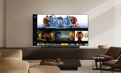 Wider view of a modern living room with a wall-mounted TV displaying films like GOAT and Anaconda. A slim soundbar sits below the screen, with a beige sofa, coffee table, and chairs completing the setup.