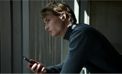 Một chàng trai trẻ đeo tai nghe màu đen vừa sử dụng điện thoại vừa nhìn ra ngoài cửa sổ.