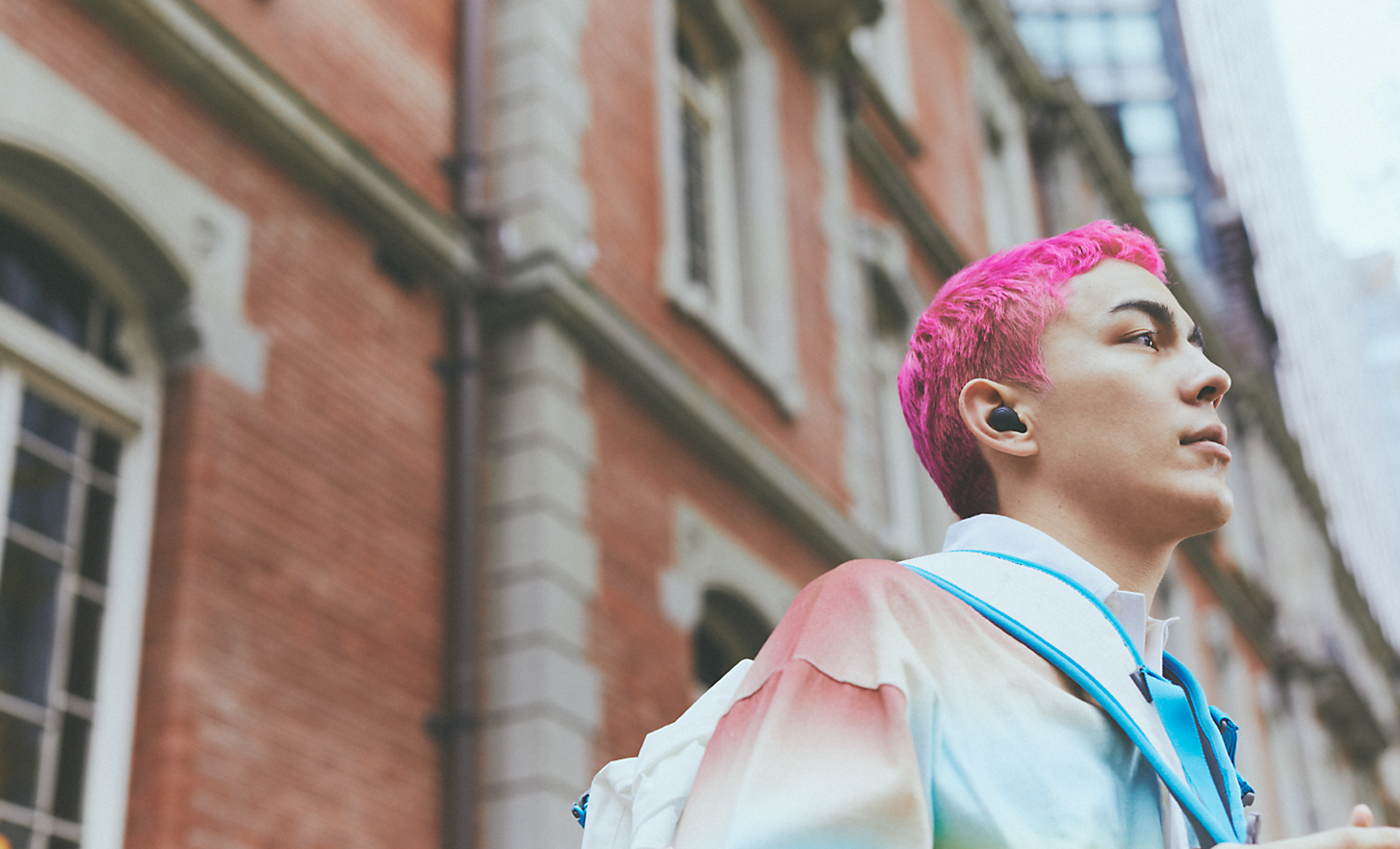 Image of a person with pink hair wearing black WF-C700N Wireless Noise Cancelling headphones in front of a brick building