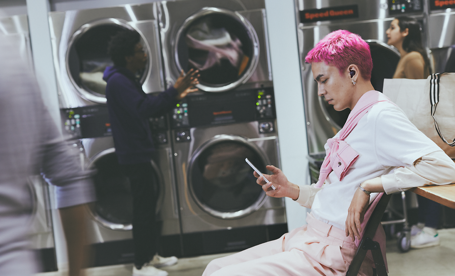 Bilde av en person på et vaskeri som ser på telefonen sin, iført trådløse WF-C700N-hodetelefoner med støyreduksjon