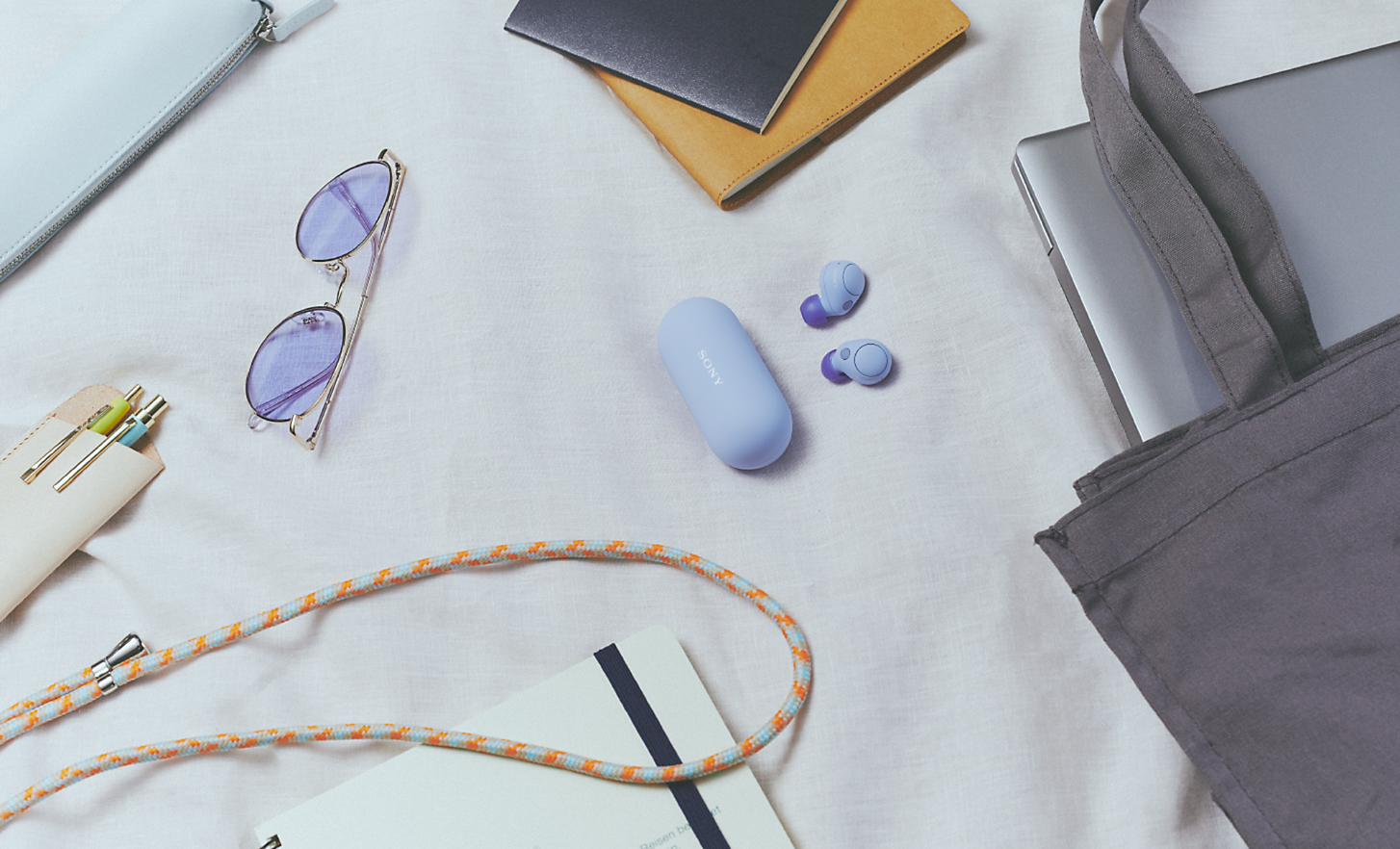 Imagem dos auriculares WF-C700N e estojo lilases, rodeados por vários itens do dia-a-dia, tais como um portátil, uma bolsa e óculos de sol