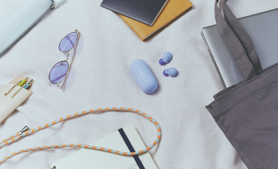 Изображение лавандовых наушников WF-C700N и чехла рядом с разнообразными предметами для повседневного использования, такими как ноутбук, сумка и солнцезащитные очки