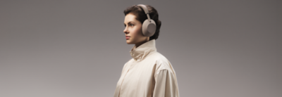 En kvinna i en beige jacka med hög krage och WH-1000XM6-hörlurar från Sony på sig, bild i profil från sidan, neutral bakgrund.