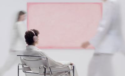 샌드 핑크 헤드폰을 착용한 젊은 남성이 카메라를 등지고 분홍색 보드 앞의 의자에 앉아 있습니다. 그의 주변에서 걸어 다니는 사람들은 흐릿하게 처리되어 있습니다.