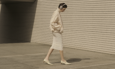 Full-length side view of a person wearing Sony WH-1000XM6 Sandstone headphones while walking past a modern, ribbed concrete wall.