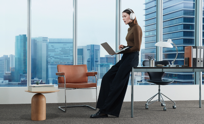 A person wearing a pair of white 1000X THE COLLEXION headphones in an office leaning against a desk holding a laptop