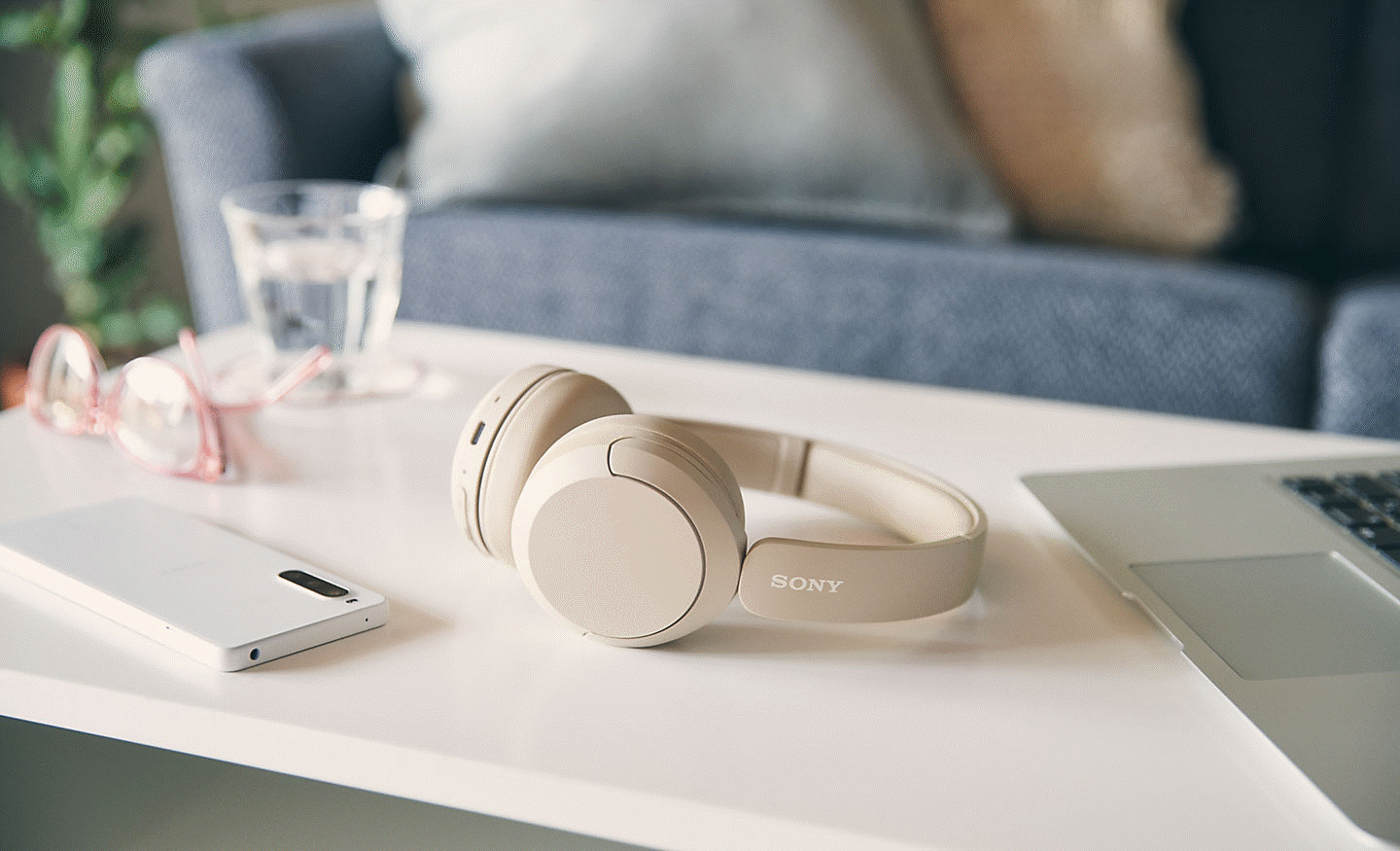 Image of a beige pair of Sony WH-CH520 headphones sitting on a coffee table with a mobile phone and laptop