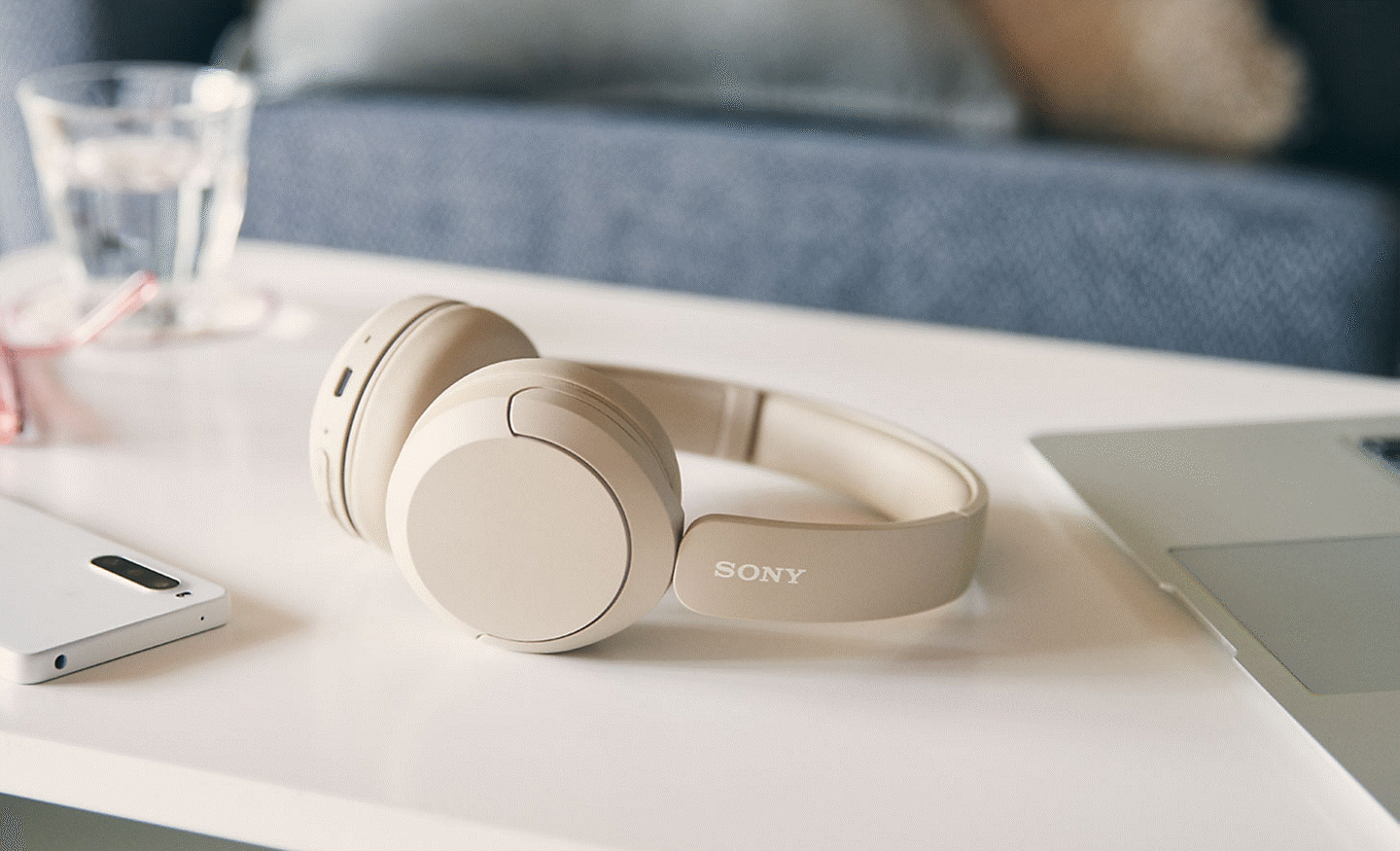 Image of a beige pair of Sony WH-CH520 headphones sitting on a coffee table with a mobile phone and laptop