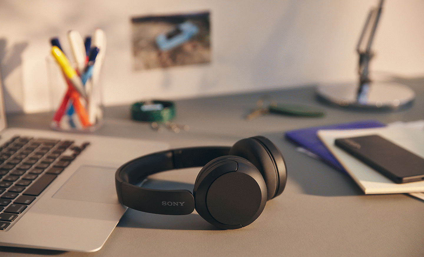 Image of a black pair of Sony WH-CH520 headphones sitting on a desk with some note books and a laptop
