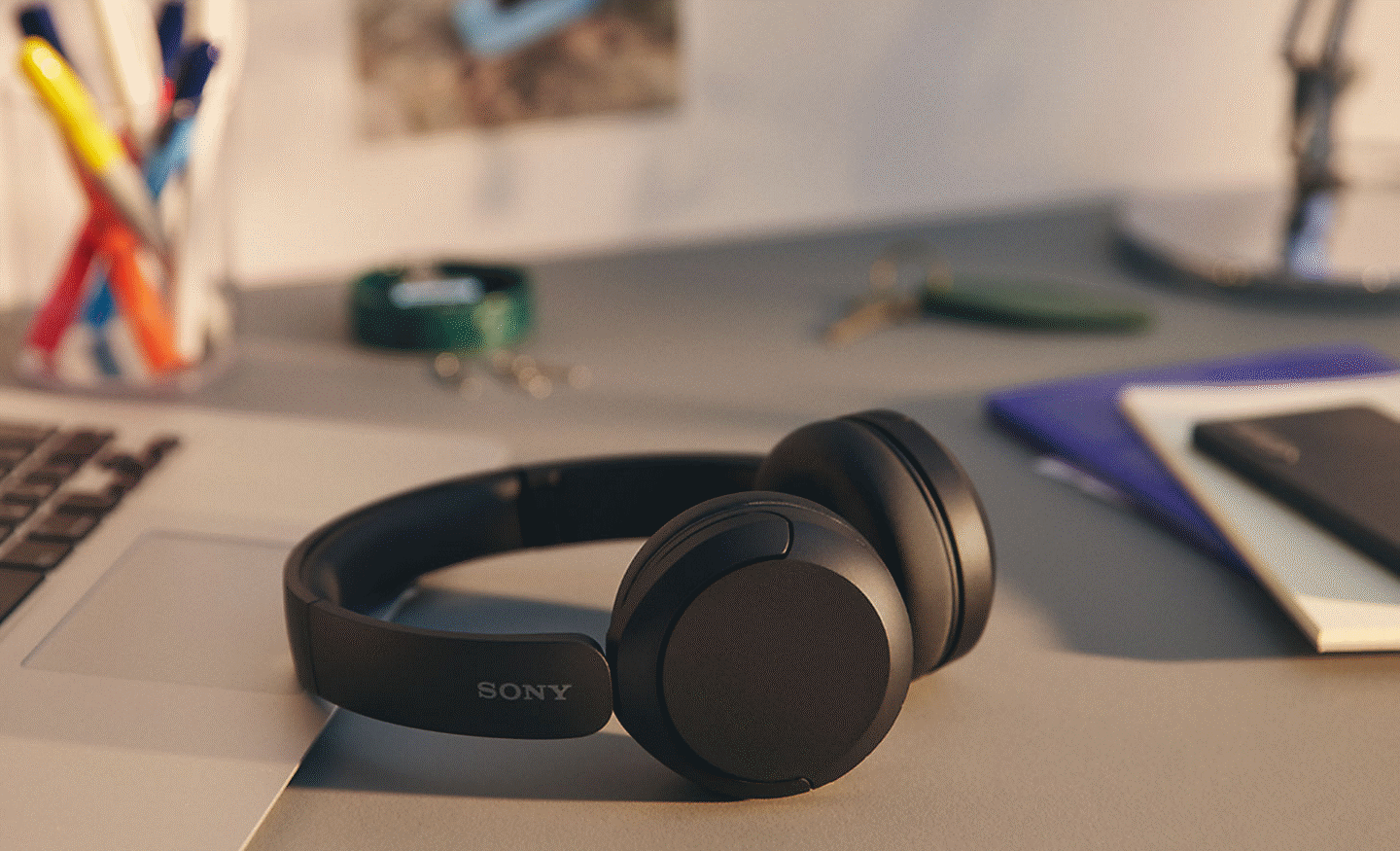 Image of a black pair of Sony WH-CH520 headphones sitting on a desk with some note books and a laptop