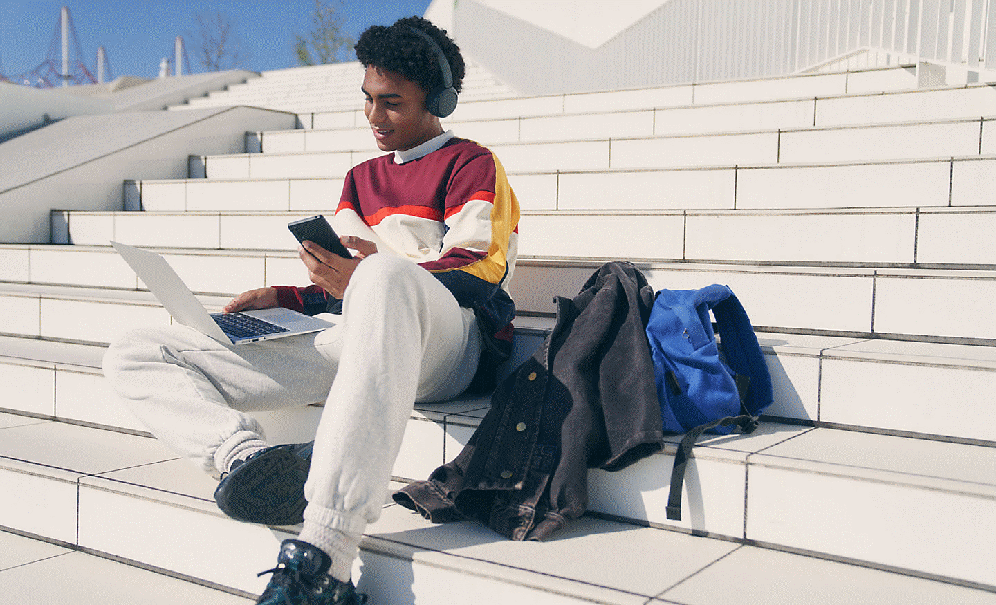 Image of a man sitting on some steps wearing Sony WH-CH520 headphones whilst using a laptop and mobile phone