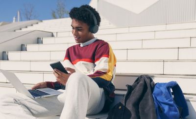 Image of a man sitting on some steps wearing Sony WH-CH520 headphones whilst using a laptop and mobile phone