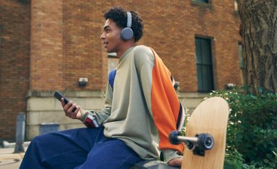 Изображение мужчины в наушниках Sony WH-CH520 и с мобильным телефоном, сидящего на стене со скейтбордом на фоне