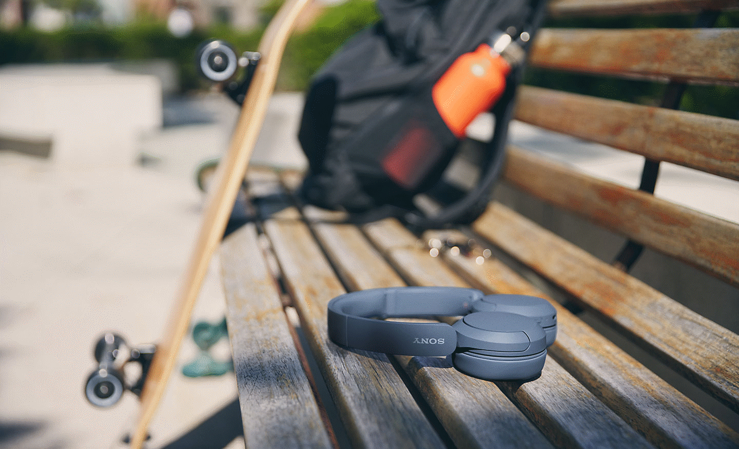 Imagem de um par de auscultadores Sony WH-CH520 pretos pousados sobre uma bancada com uma mochila e skate em fundo