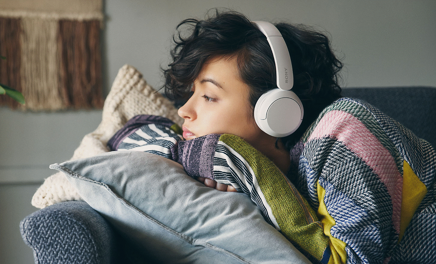 Изображение на жена, легнала върху сив диван, с чифт бели слушалки Sony WH-CH520