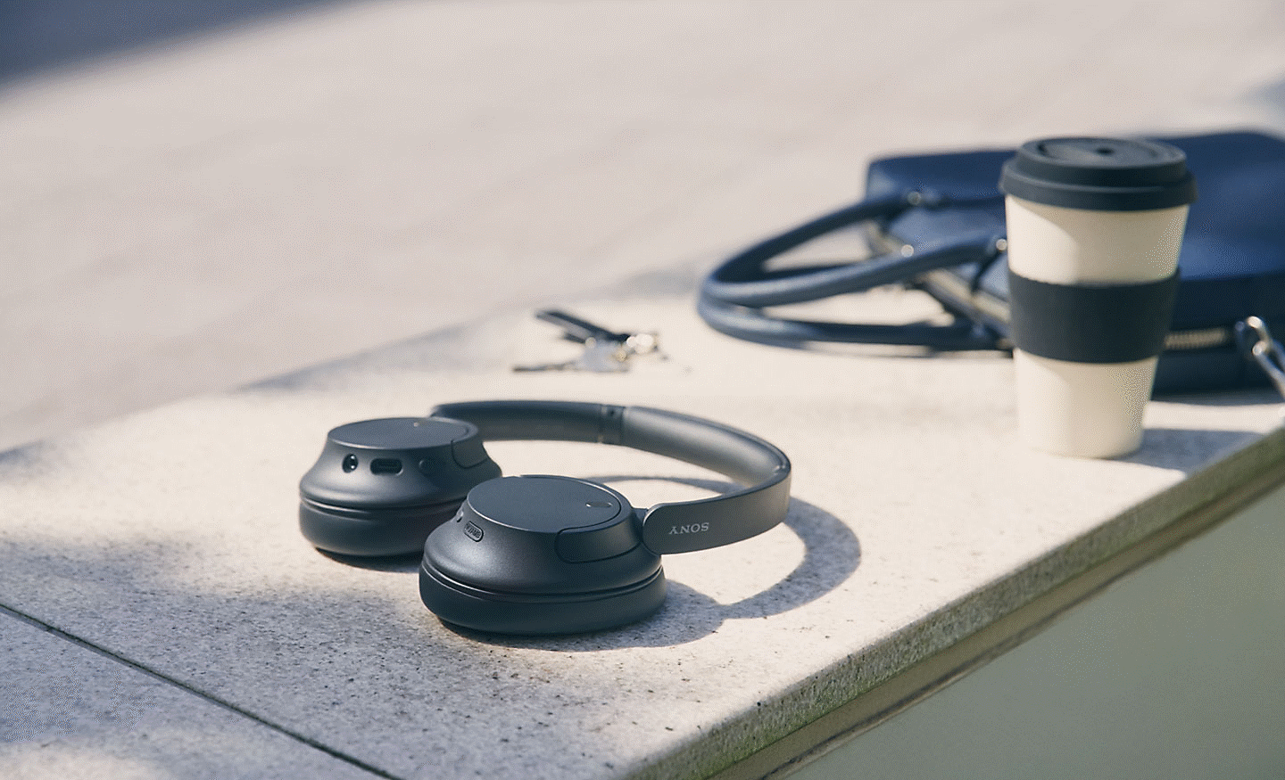 Image of a black pair of Sony WH-CH720 headphones sitting on a wall with a coffee cup and bag