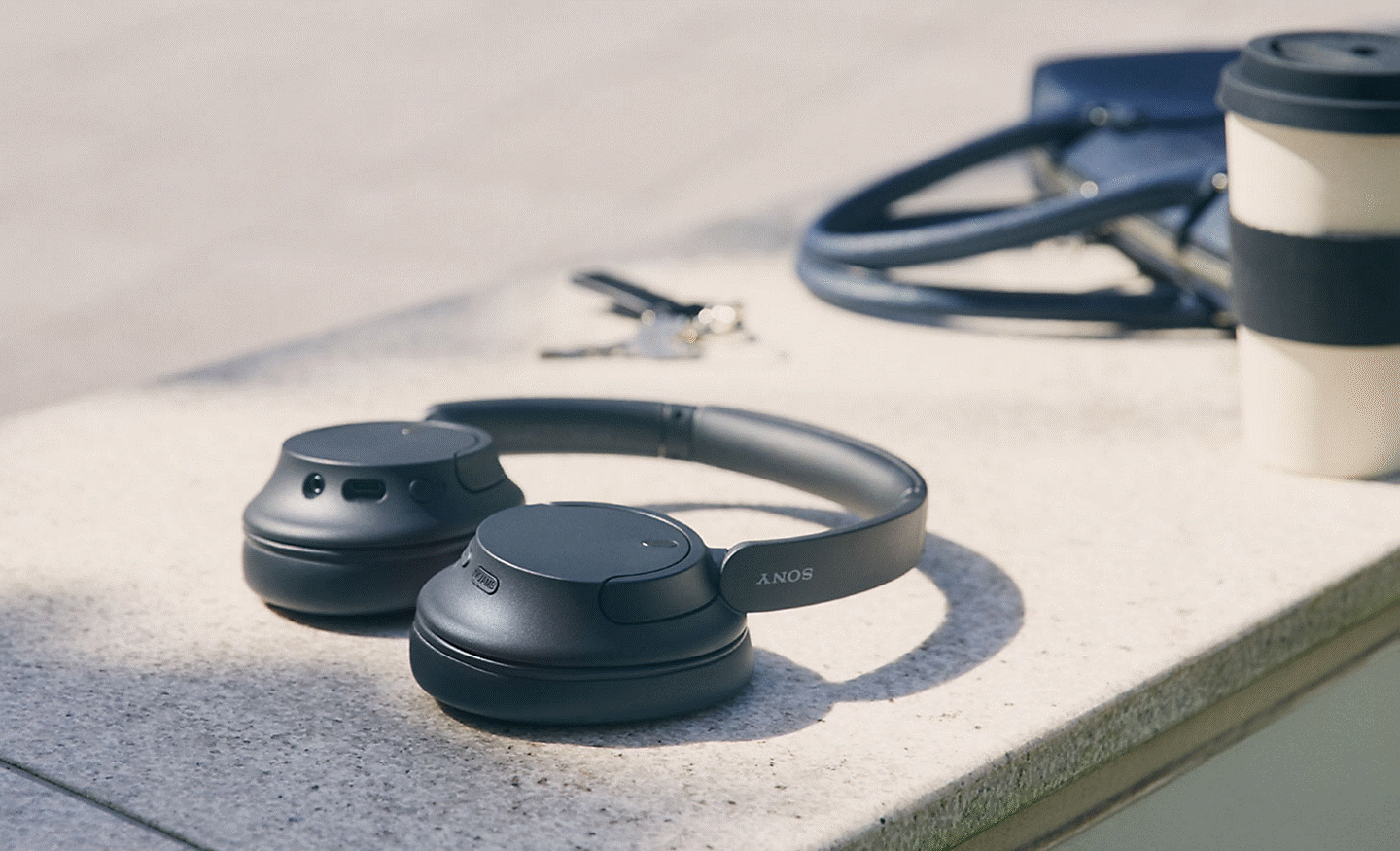 Imagen de unos audífonos negros WH-CH720 de Sony apoyados en una pared con una taza de café y una bolsa