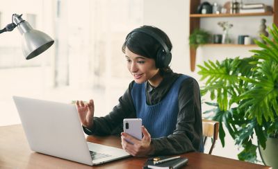 Hình ảnh một người phụ nữ ở bàn làm việc đeo cặp tai nghe Sony WH-CH720 màu đen, đang sử dụng máy tính xách tay và điện thoại di động