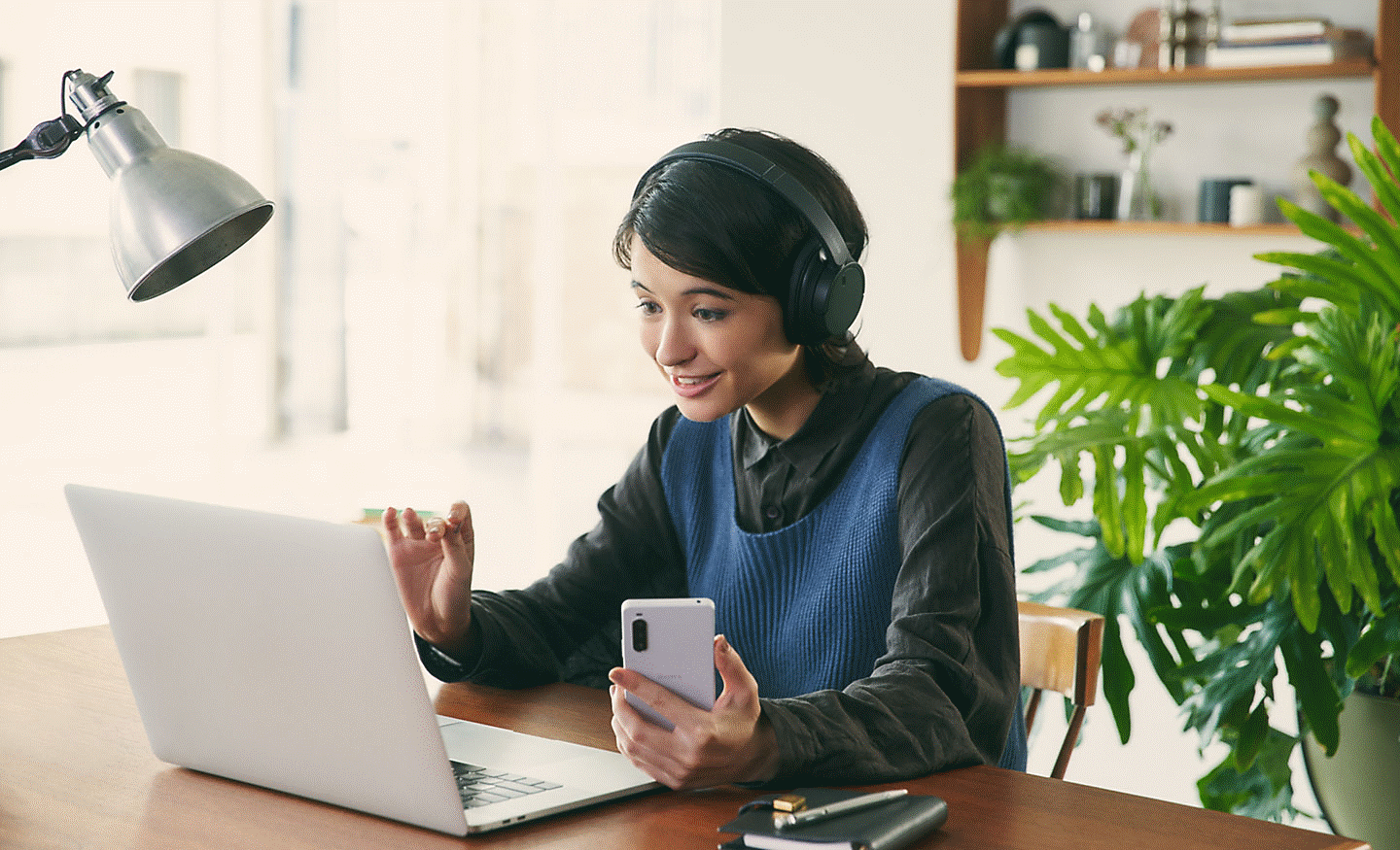 Slika ženske, ki za mizo nosi črne slušalke Sony WH-CH720 ter uporablja prenosni računalnik in mobilni telefon