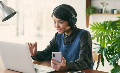 תמונה של אישה ליד שולחן מרכיבה זוג שחור של אוזניות Sony WH-CH720 ומשתמשת במחשב נייד ובטלפון נייד