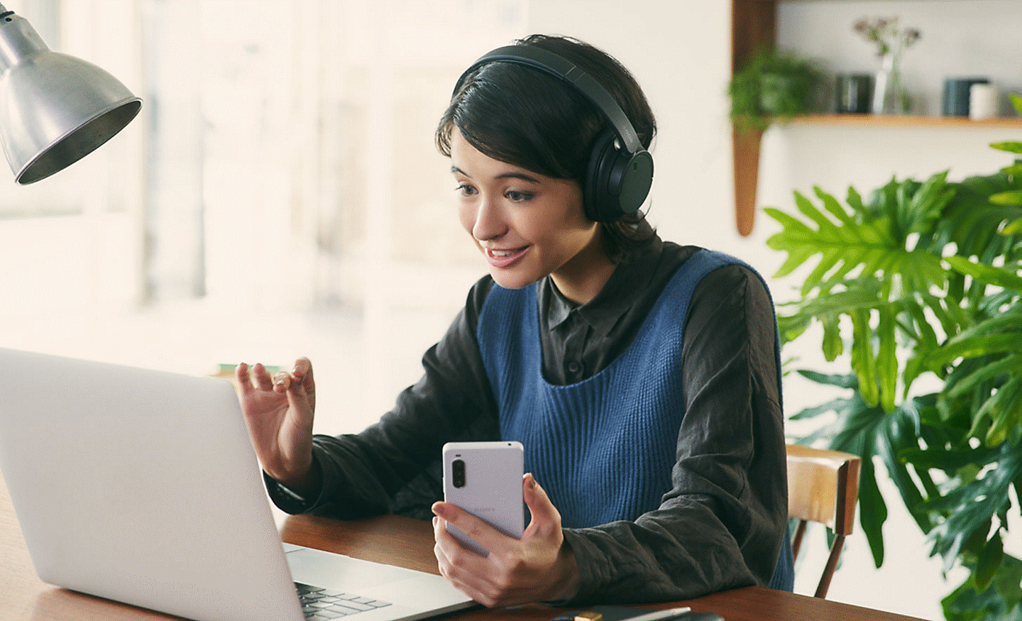 Image of a woman at a desk wearing a black pair of Sony WH-CH720 headphones and using a laptop and mobile phone