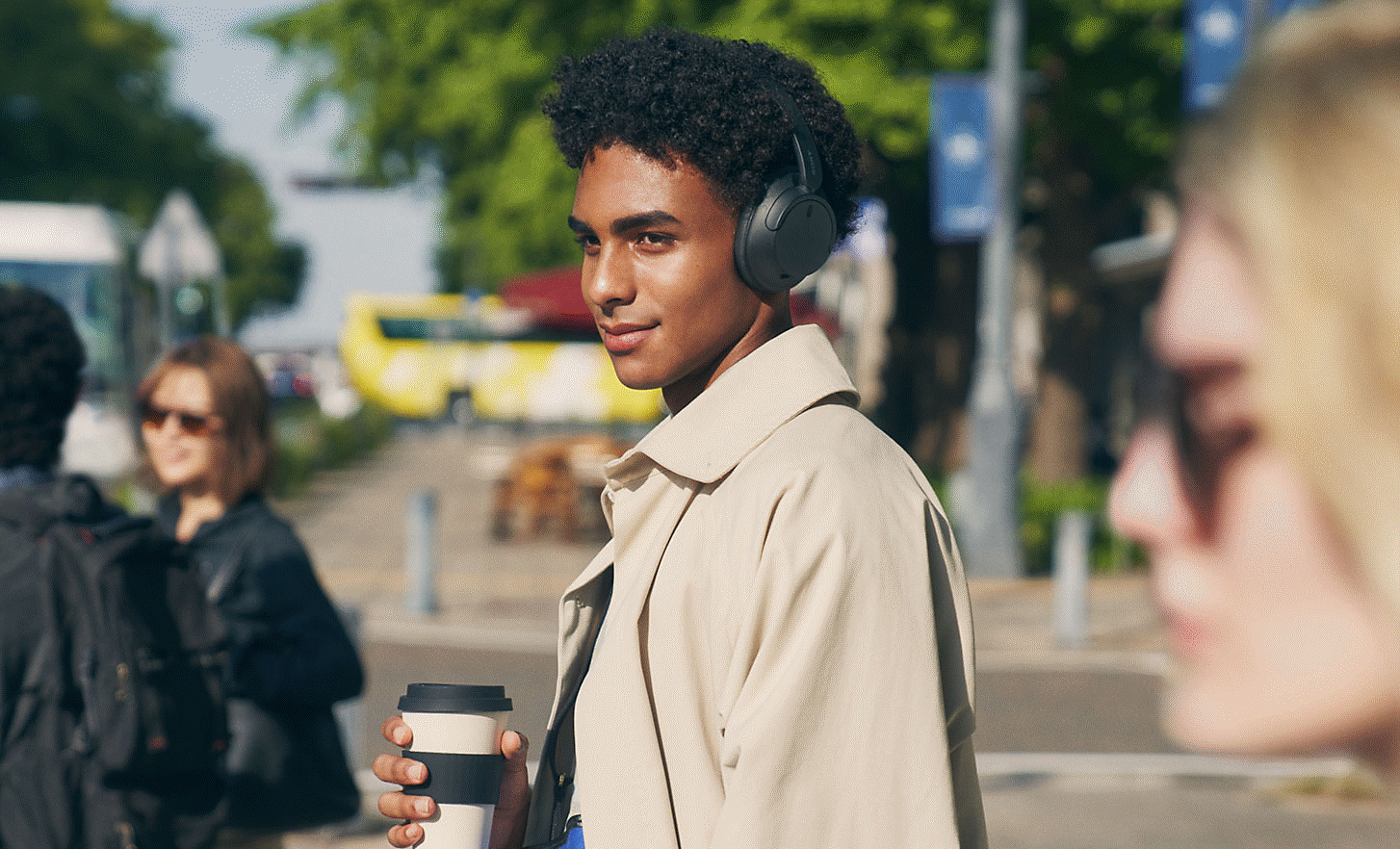 Изображение на човек в град, който носи чифт черни слушалки Sony WH-CH720 и държи чаша кафе