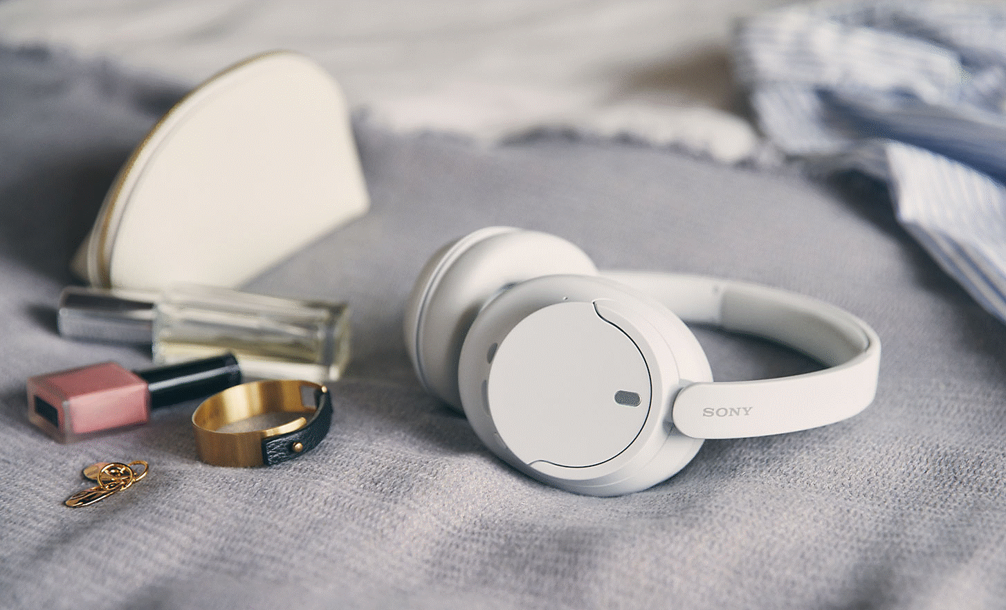 Gambar headphone putih Sony WH-CH720 di tempat tidur dengan beberapa aksesori