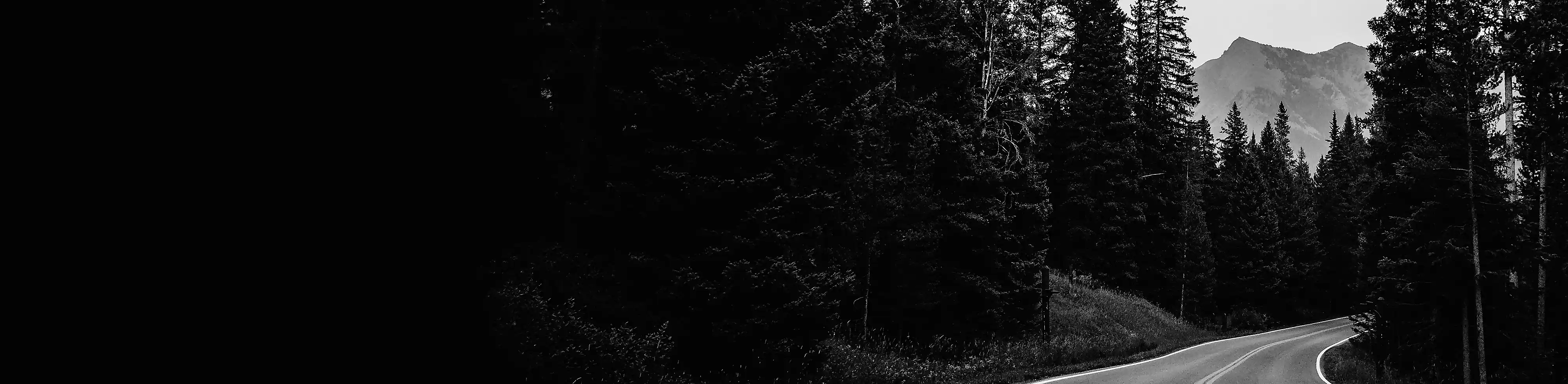 En tvåfilig väg som löper genom skogen med ett berg i bakgrunden, i svartvitt.