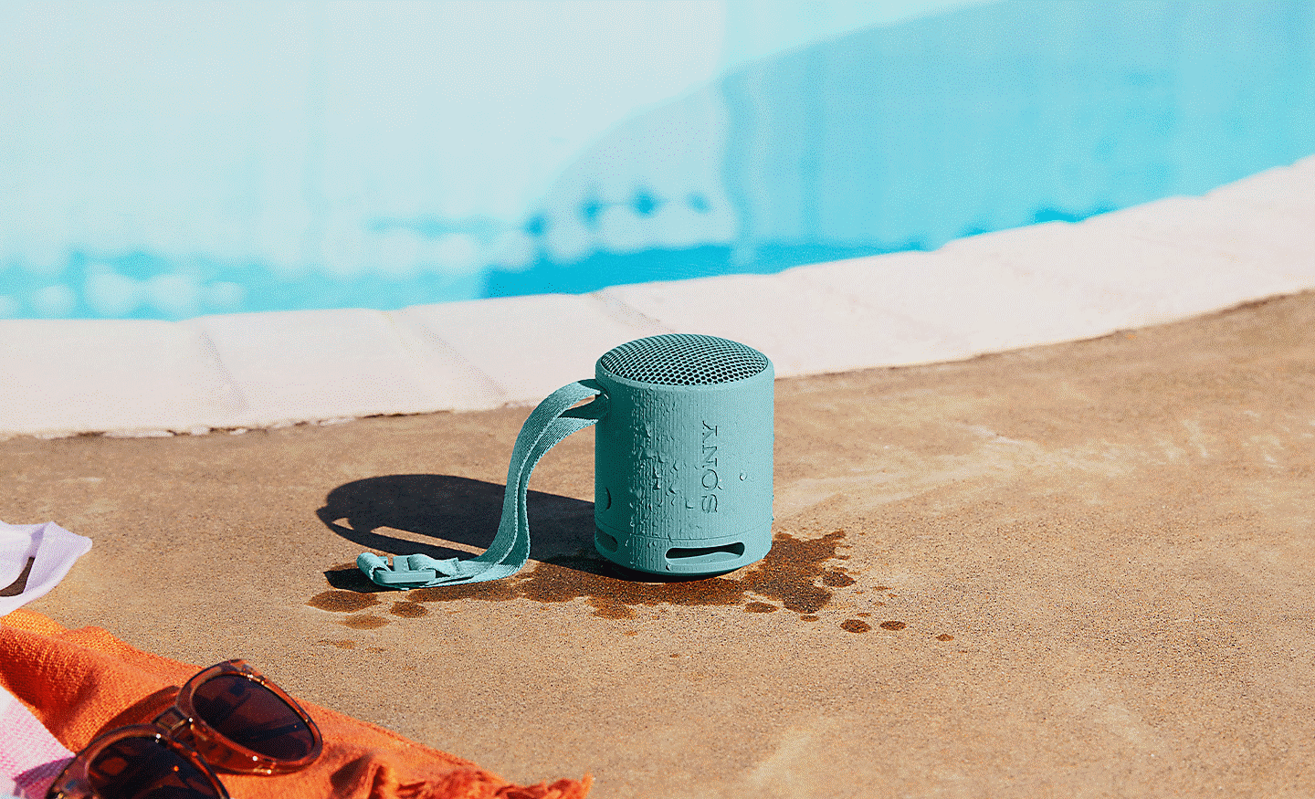 Image of a wet blue SRS-XB100 speaker sitting on the side of a swimming pool. Splash marks surround the speaker