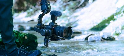 Obrázok osoby, ktorá používa kameru namontovanú na stabilizátore a nahráva zvuk blízko malej rieky.
