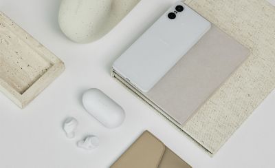 The closed white charging case shown on a desk looking down with the white ear buds and a white mobile phone