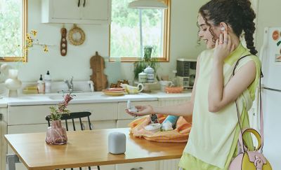 A young woman wearing LinkBuds Fit earbuds holding the charging case in her hand with a LinkBuds Speaker on a table nearby.