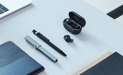 Un escritorio de trabajo con el estuche de carga negro abierto que contiene un audífono negro y el otro audífono negro colocado al lado en el escritorio