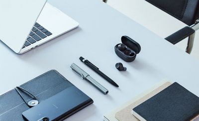 مكتب عمل مع علبة شحن سوداء مفتوحة عليه تحتوي على سماعة أذن سوداء واحدة مع سماعة أذن سوداء أخرى موضوعة بجانبها على المكتب
