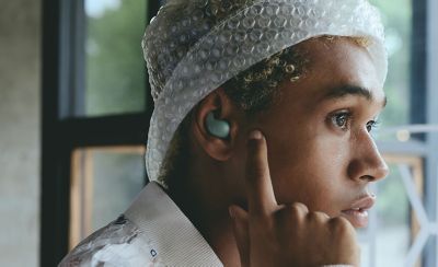 A young man wearing a hat adjusting the audio on LinkBuds Fit earbud by tapping the side of his face next to his ear.