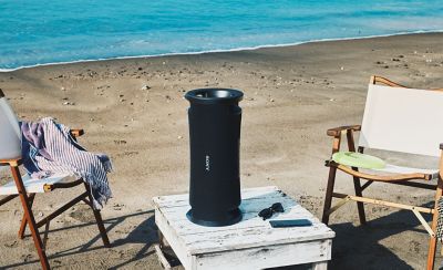 A beach with the ULT FIELD 7 portable speaker on a table beside two deck chairs.