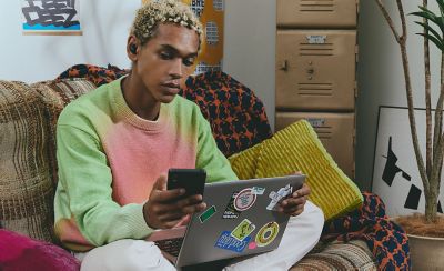 A young man wearing LinkBuds Fit sat on a couch holding his smartphone with his laptop on his lap.