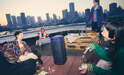  Four people sit on a roof top with a city skyline in the background around an SRS-XV800 speaker with purple ambient lighting
