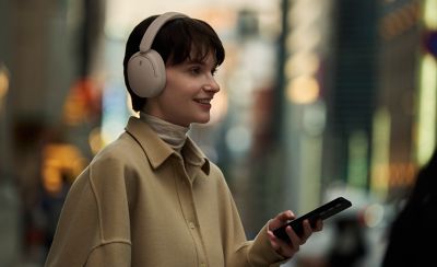 O femeie purtând o pereche de căști Sony WH-CH730N bej, ținând un smartphone pe o stradă